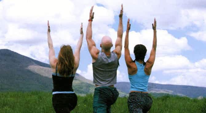 Three people doing yoga in a field with mountains in the background.