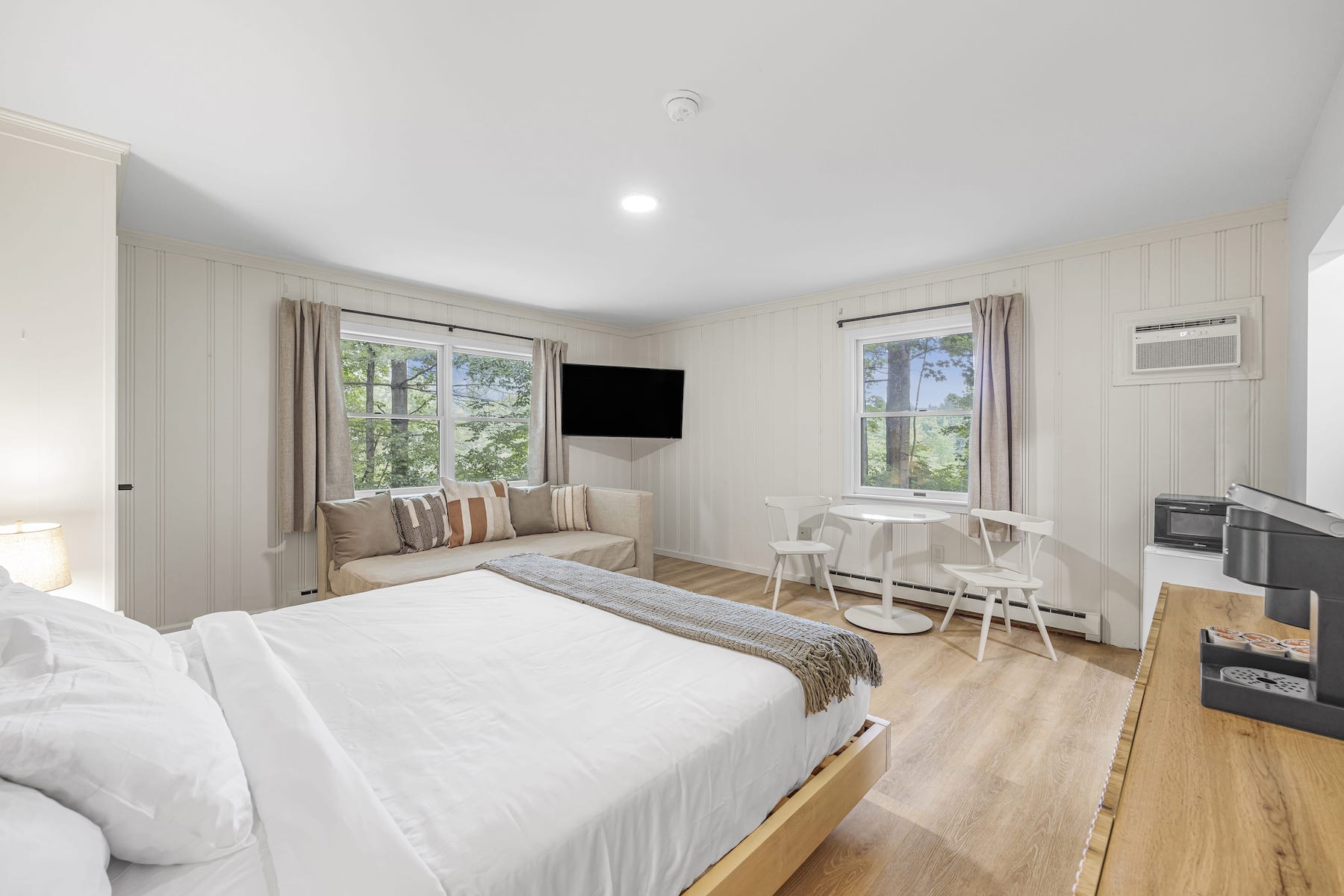 Cozy bedroom with a double bed, white linens, a sofa, TV, small dining table with chairs, and two windows showcasing a leafy outdoor view.