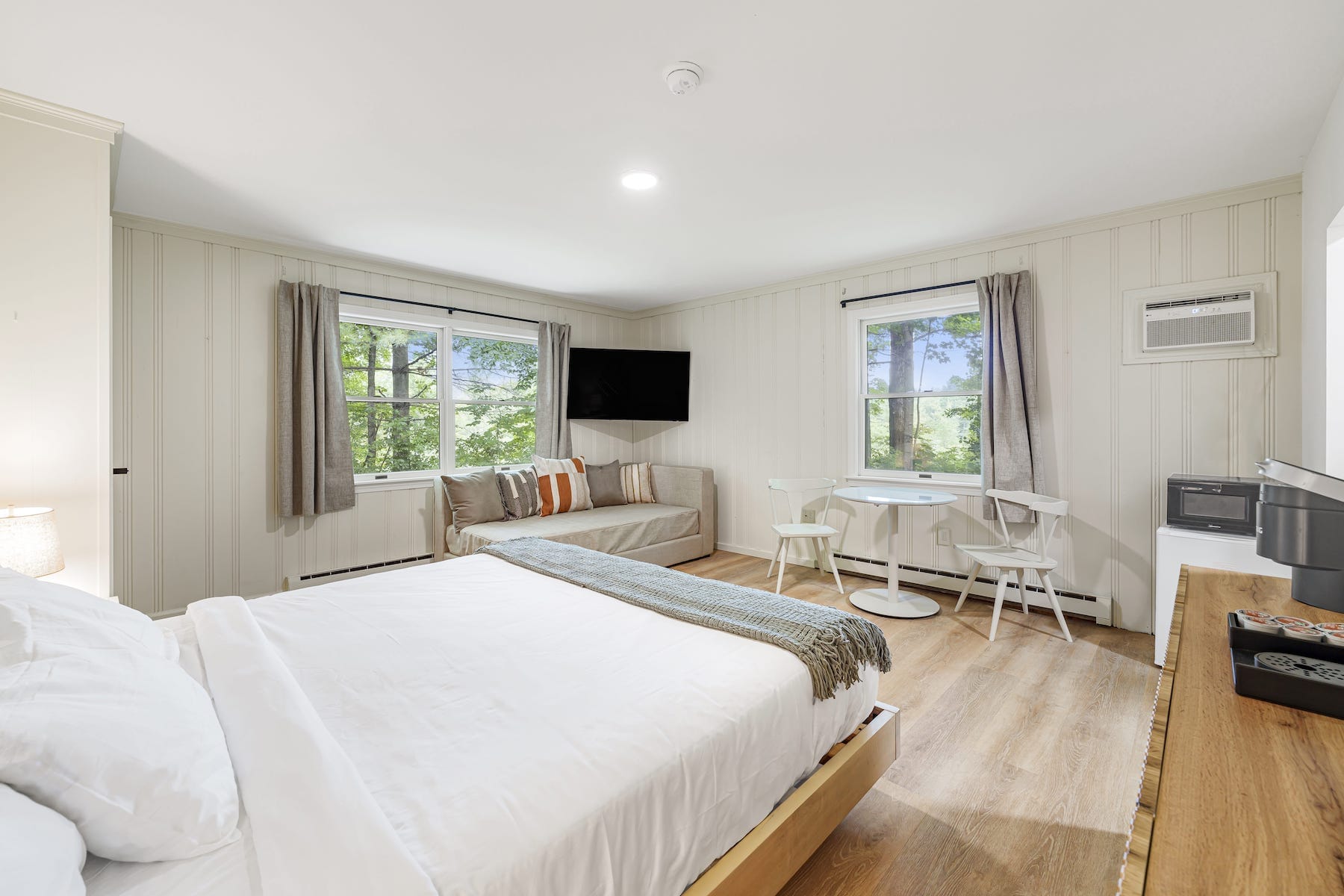 Spacious, bright bedroom with a large bed, wooden floors, windows, a couch, a wall-mounted TV, and a small dining area with a round table and two chairs.