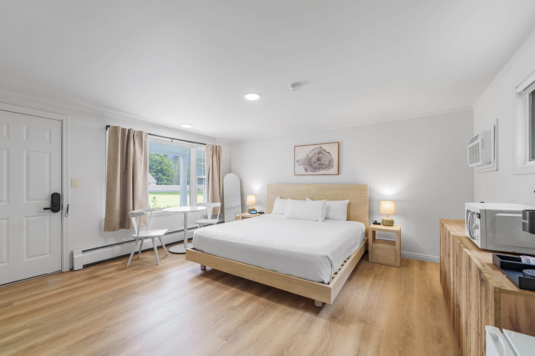 Minimalist hotel room with a large bed, wooden furniture, and natural light from a window. Includes a small dining area, microwave, and wall art. Wood flooring and neutral tones throughout.