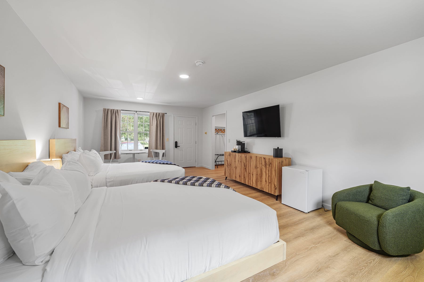 A hotel room with two double beds, a flat-screen TV, a wooden dresser, a mini fridge, and a green armchair. There's a window with curtains and a door leading outside.