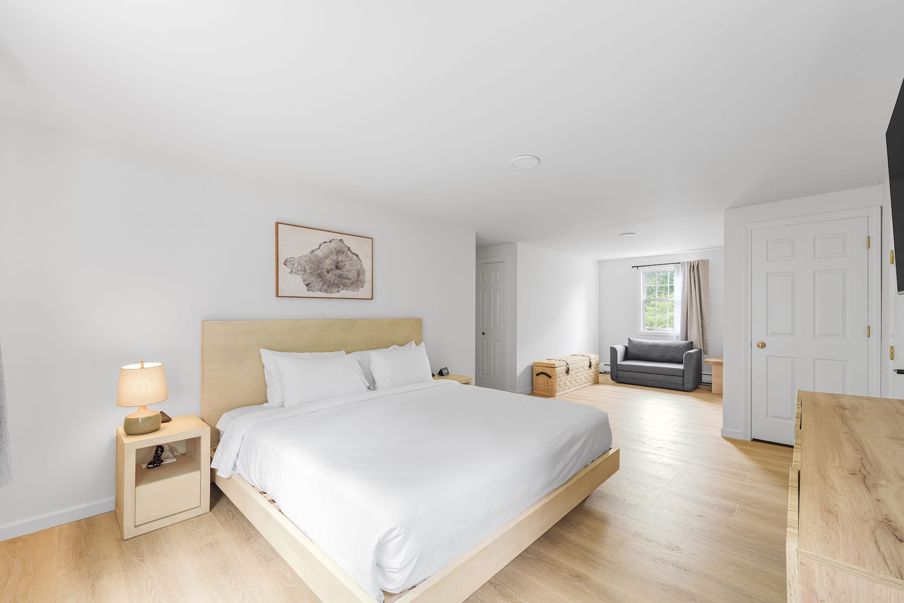 Spacious bedroom with a wooden bed, white bedding, a bedside lamp, and a dresser. A separate area features a gray sofa, basket, and window with curtains. Walls are white, and the floor is wooden.