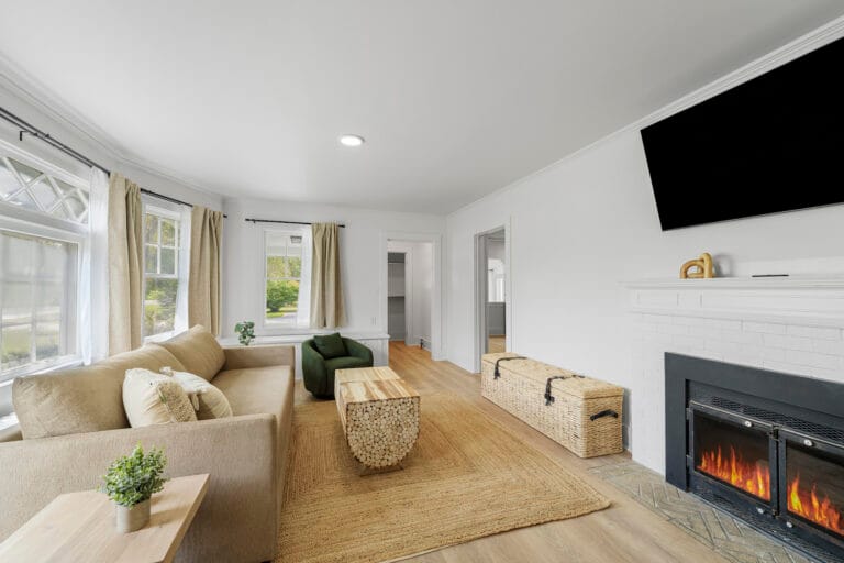 A living room with a sofa, armchair, wooden coffee table, fireplace, and wall-mounted TV. Large windows with curtains provide natural light. A woven basket is placed in front of the fireplace.