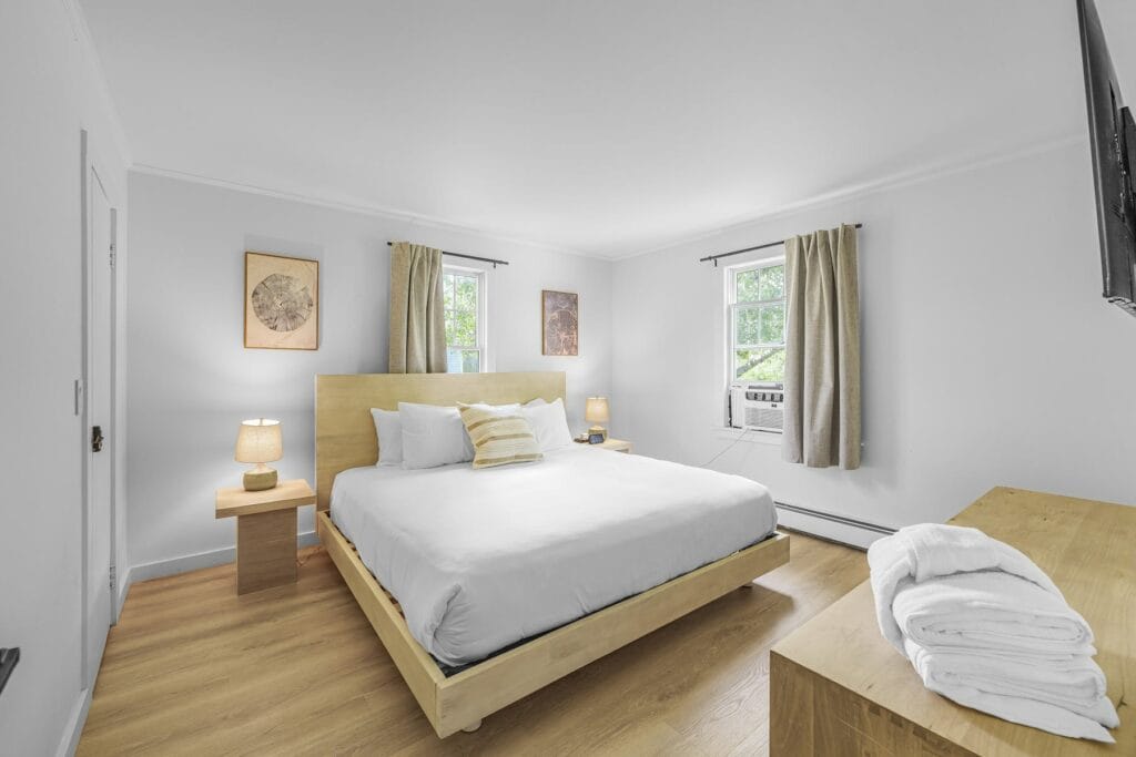 Modern bedroom with a large bed, wooden furniture, two windows, and neatly folded towels on a dresser.