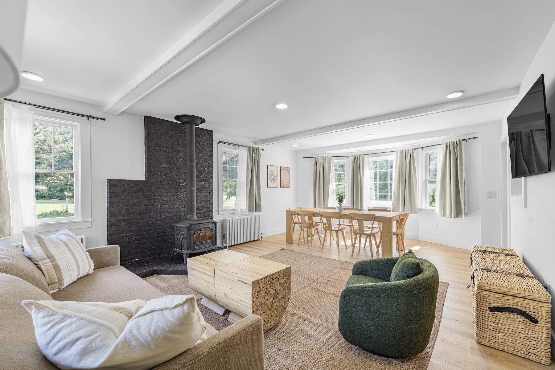 A bright living room with a beige sofa, green armchair, wooden tables, black fireplace, large windows with curtains, a wall-mounted TV, and a dining table with chairs on a woven rug.