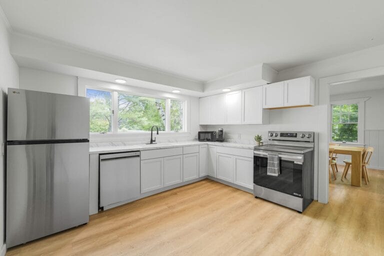 A modern kitchen with white cabinets, stainless steel appliances, a large window, and wooden flooring. A dining area is visible in the background.