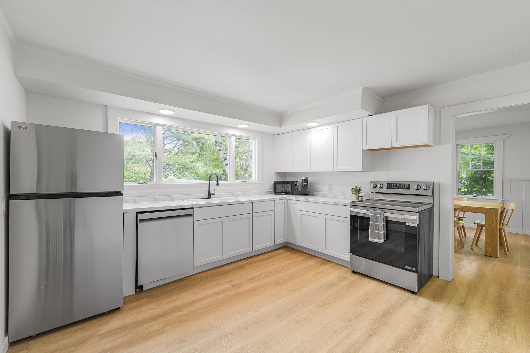 A modern kitchen with white cabinets, stainless steel appliances, a large window, and wooden flooring. A dining area is visible in the background.