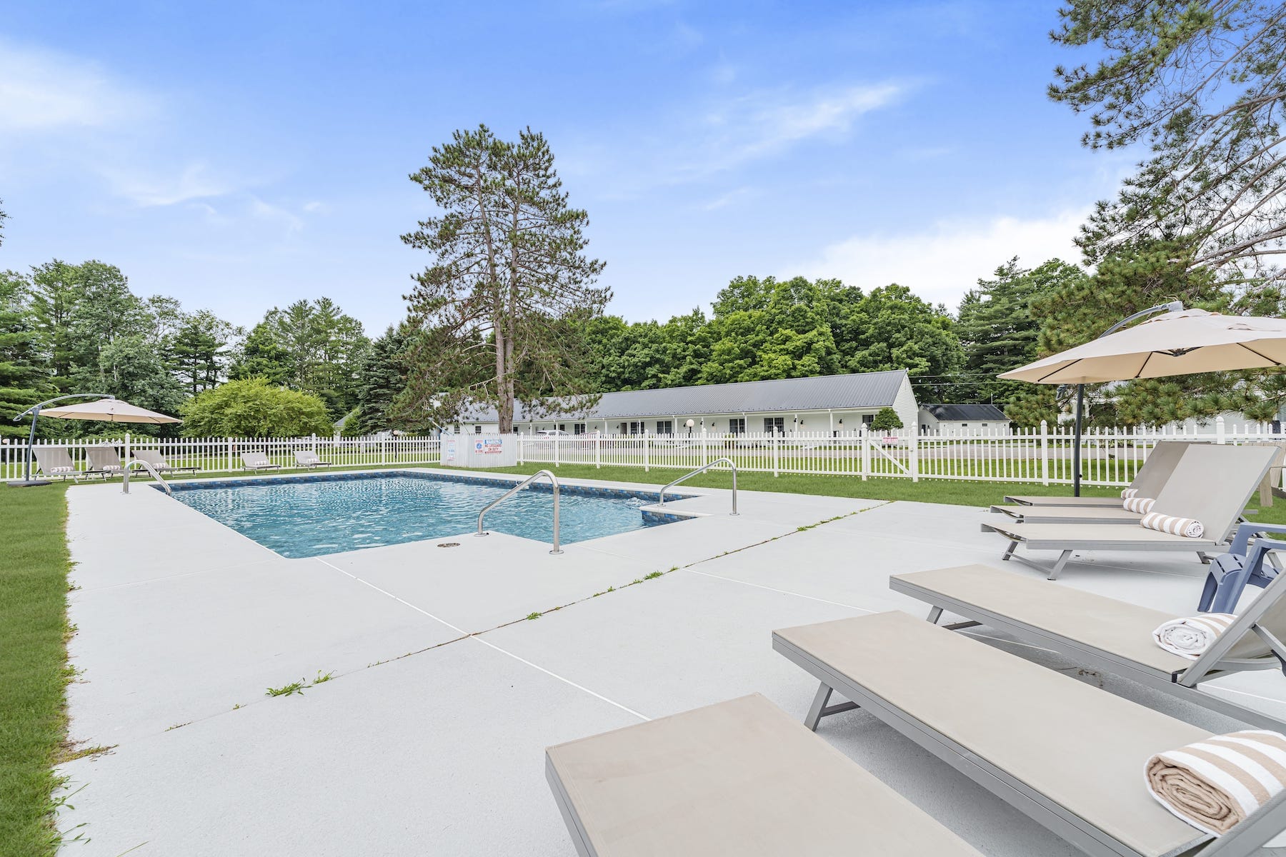 Outdoor swimming pool with white lounge chairs, umbrellas, and towels on a concrete deck, surrounded by grass, trees, and a white picket fence under a blue sky.
