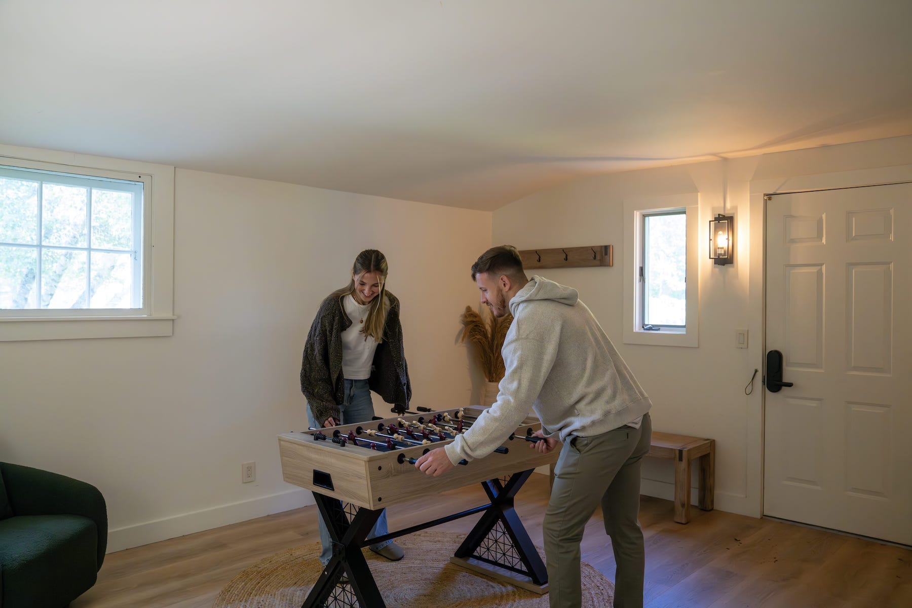 Two people play foosball in a well-lit room with a door and windows. The room has light-colored walls and wood flooring.