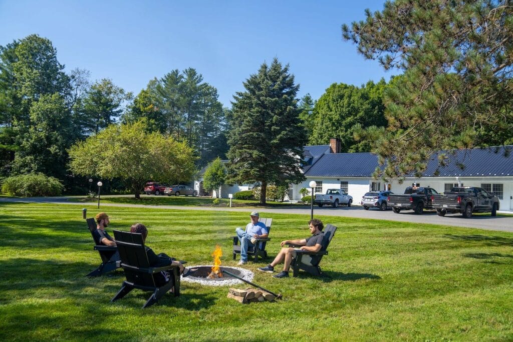 Four people sit in chairs around a small fire pit on a grassy lawn with trees and a building in the background.