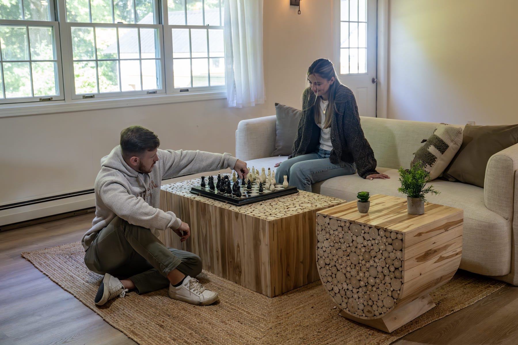 A man and woman play chess together in a living room, with natural light coming through large windows behind them.