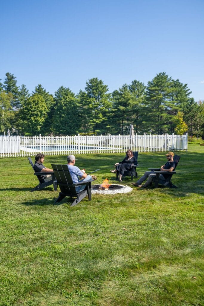 Four people sit in Adirondack chairs around a fire pit on a grassy lawn, surrounded by trees and a white fence under a clear blue sky.