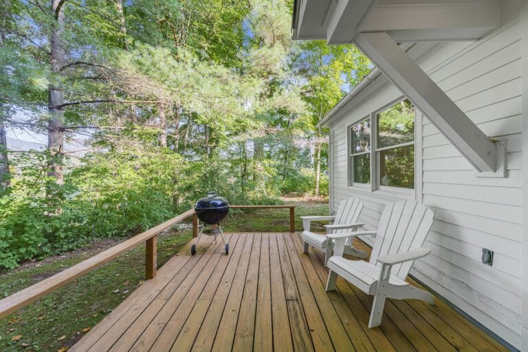 Wooden deck with two white chairs, a charcoal grill, and surrounded by trees.