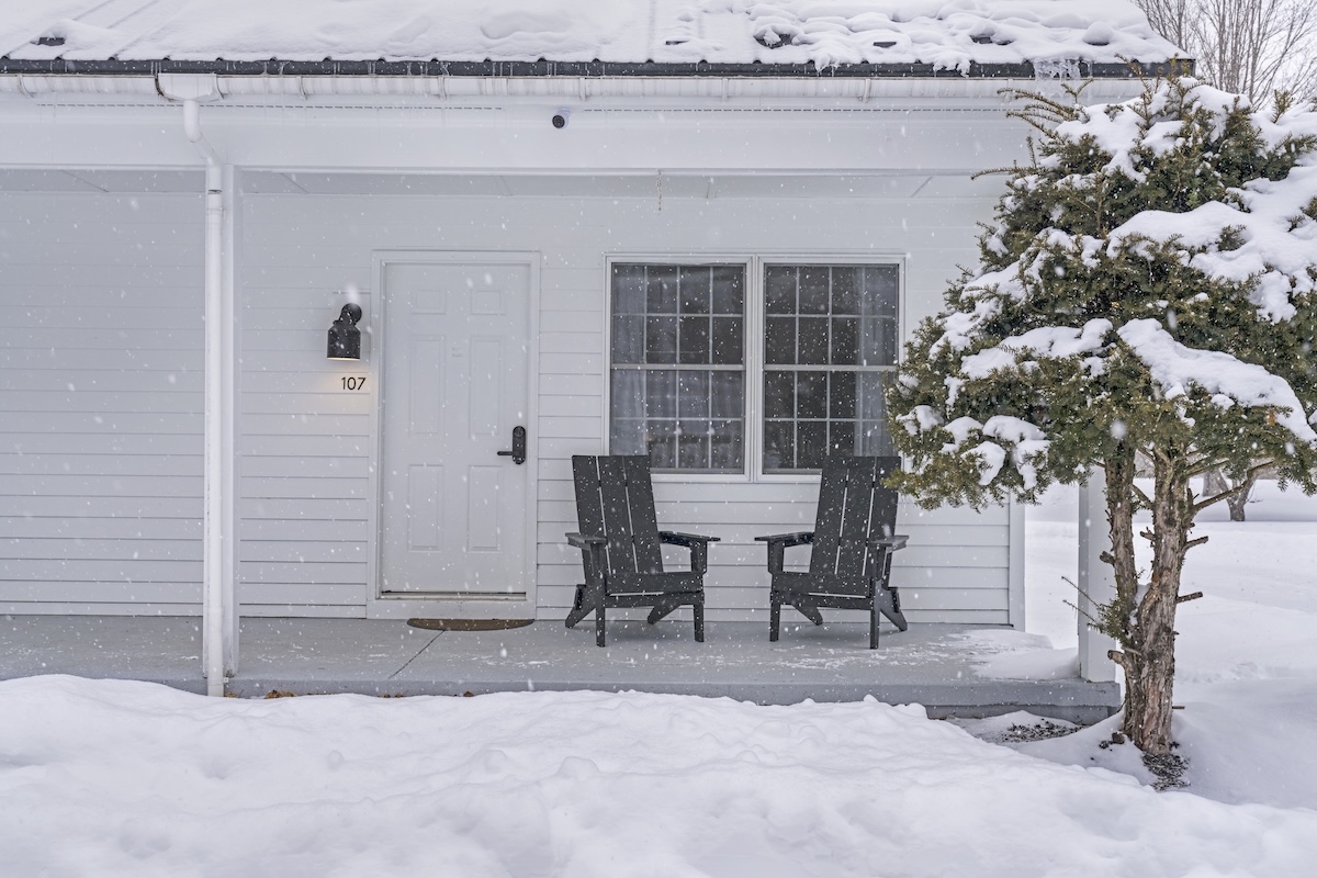 Snow-covered porch with two black chairs, a small evergreen tree, and a white door numbered 107. Snow is actively falling, covering the ground and roof.