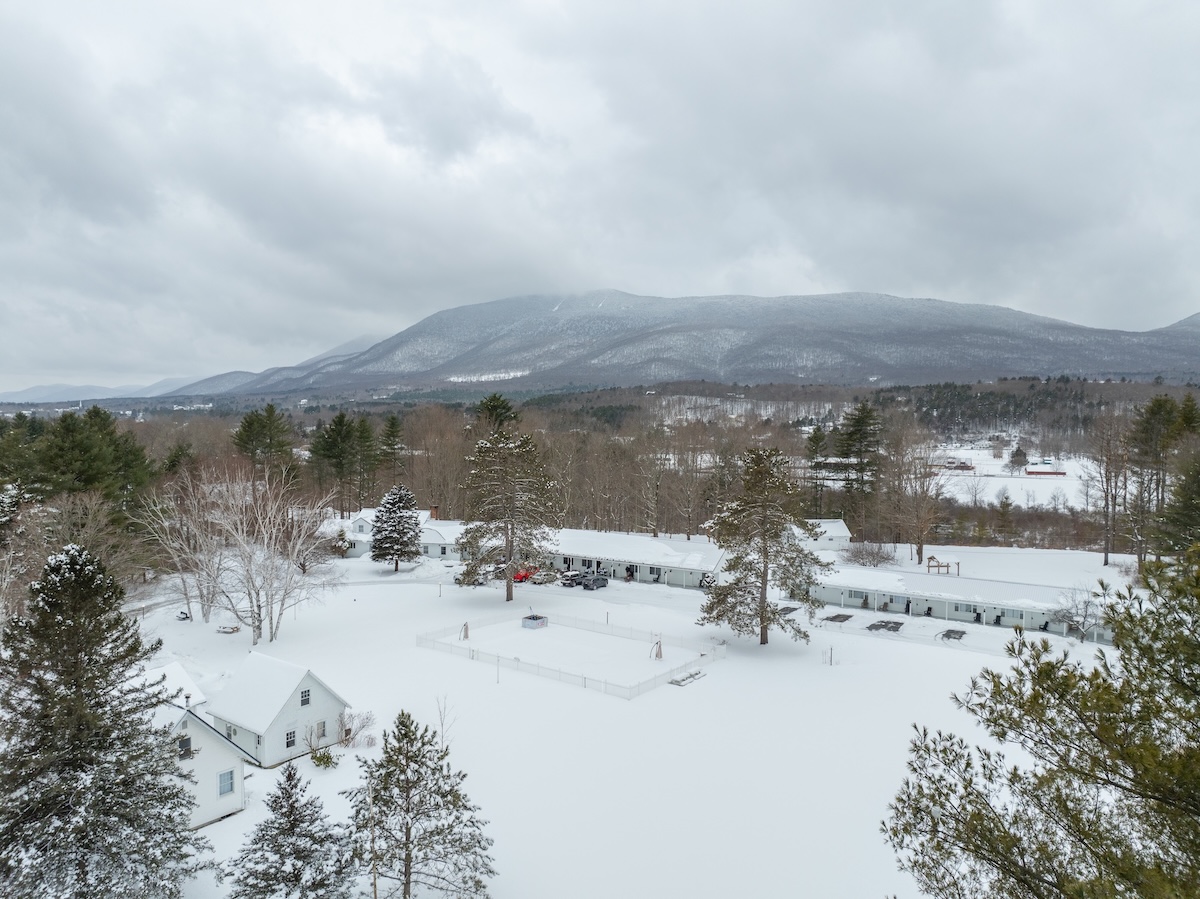 A snowy landscape with houses and trees, surrounded by mountains under a cloudy sky.