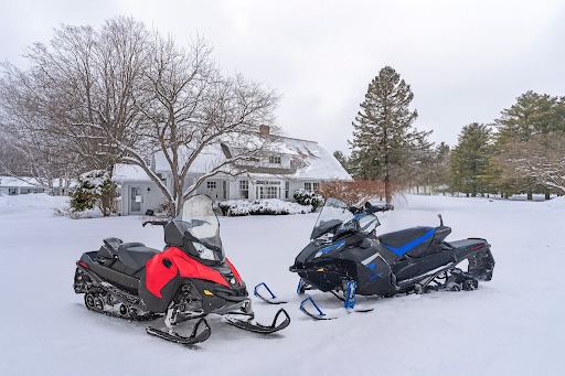 Two snowmobiles, one red and one black with blue accents, parked on snow in front of a house surrounded by trees in winter.